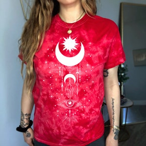 May include: Red tie-dye t-shirt with a white sun, moon, and eye design. The design also includes stars, crystals, and a crescent moon.
