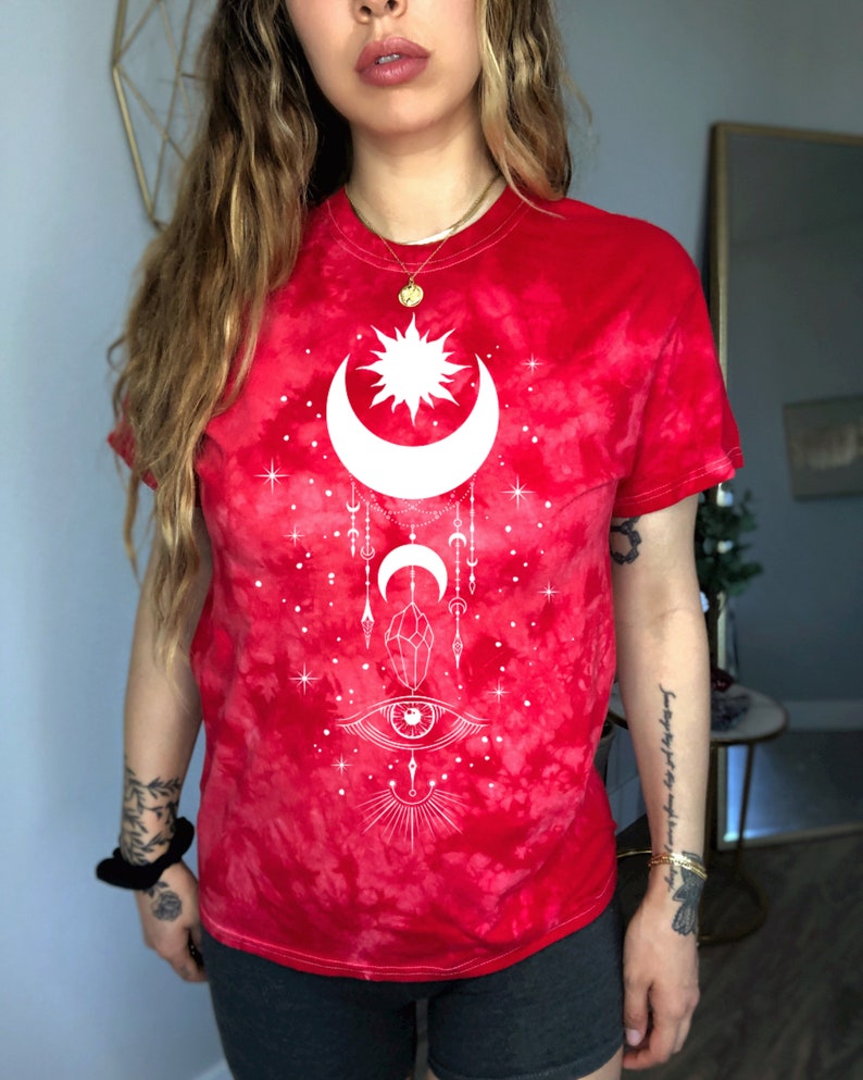 May include: Red tie-dye t-shirt with a white sun, moon, and eye design. The design also includes stars, crystals, and a crescent moon.