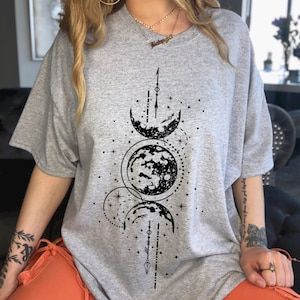 May include: A gray t-shirt featuring a black celestial design with phases of the moon and stars. The graphic is centered on the front of the shirt. The shirt has short sleeves and a relaxed fit.