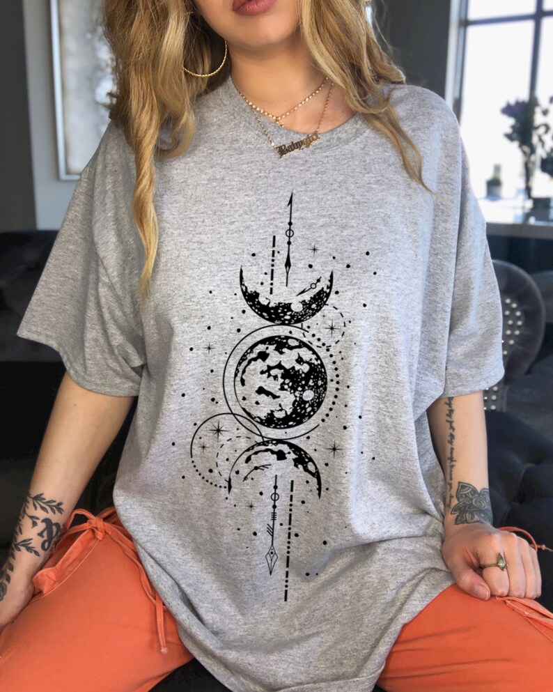 May include: A gray t-shirt featuring a black celestial design with phases of the moon and stars. The graphic is centered on the front of the shirt. The shirt has short sleeves and a relaxed fit.