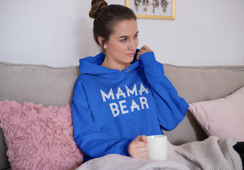 May include: A woman wearing a royal blue hoodie with the text "MAMA BEAR" in white on the front. She is sitting on a couch with a pink pillow and a white mug in her hand.