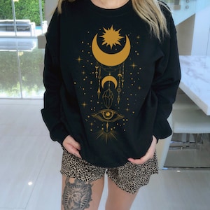 May include: Black sweatshirt with a gold crescent moon, sun, and eye design. The design also includes stars, crystals, and other celestial elements.