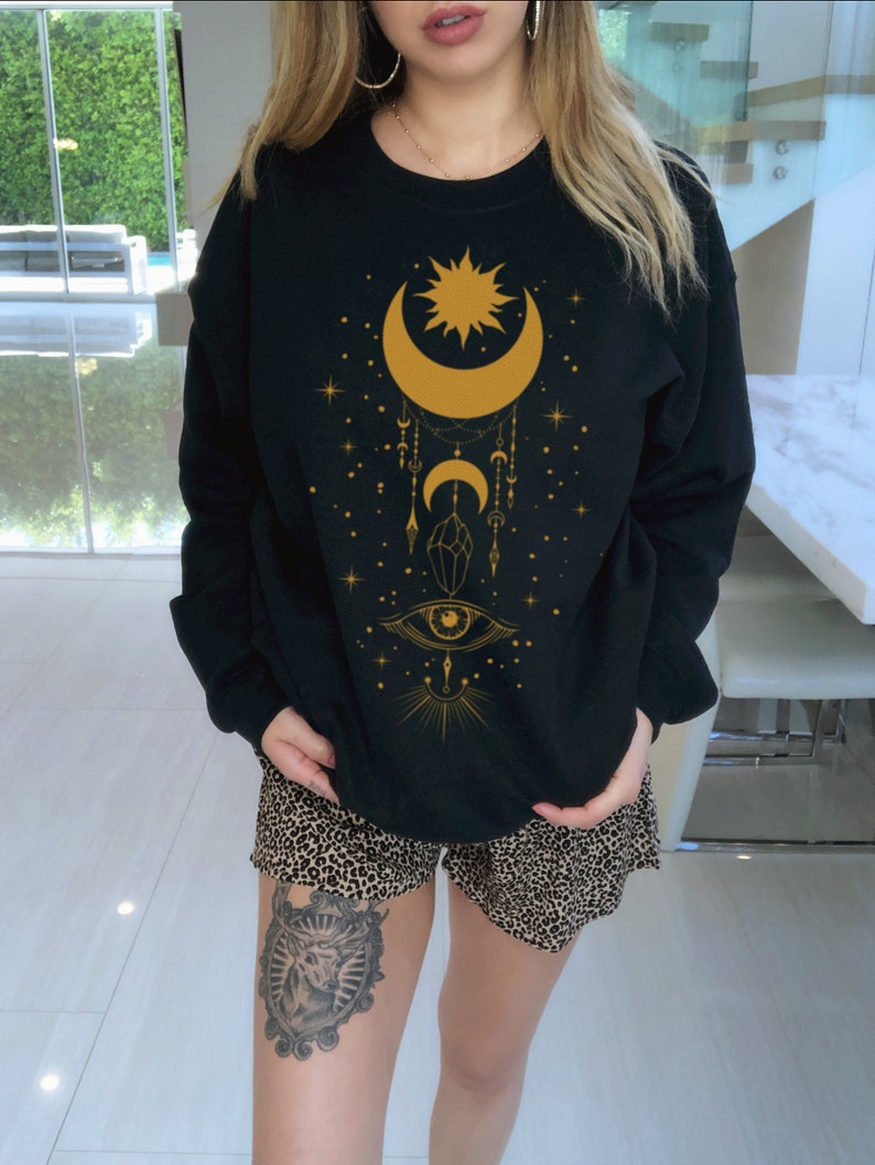 May include: Black sweatshirt with a gold crescent moon, sun, and eye design. The design also includes stars, crystals, and other celestial elements.