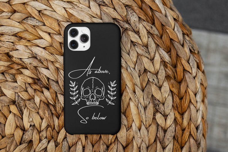Op de afbeelding: Zwarte telefoonhoes met een witte schedel en bloemenontwerp. De tekst "As above, so below" staat op de hoes.