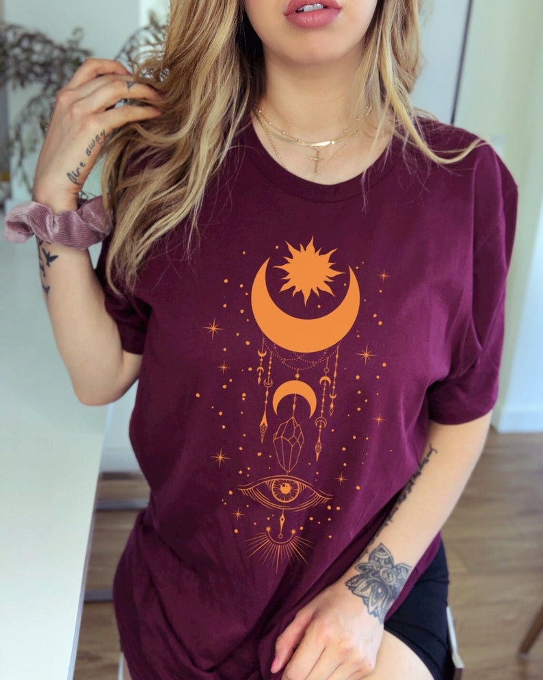 May include: Burgundy t-shirt with a graphic design of a crescent moon, sun, stars, and an eye with a crystal hanging from it.