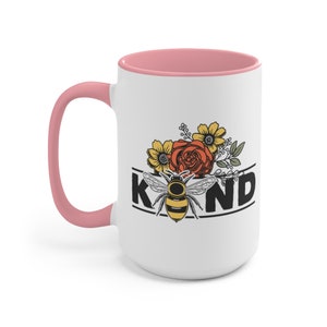 Op de afbeelding: Witte keramische mok met een roze rand en handvat. De mok heeft een zwart-witte afbeelding van een bij met bloemen en het woord "KIND".