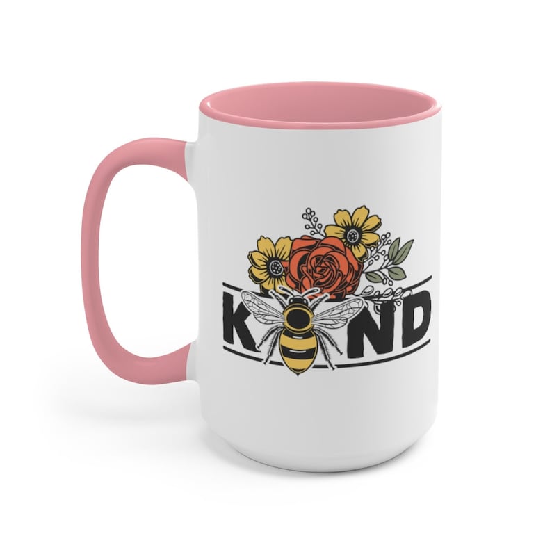 Op de afbeelding: Witte keramische mok met een roze rand en handvat. De mok heeft een zwart-witte afbeelding van een bij met bloemen en het woord "KIND".