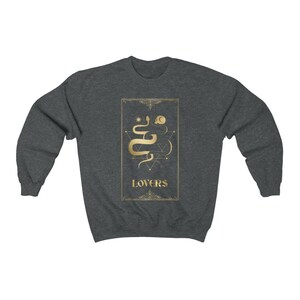 Puede incluir: Una sudadera gris oscuro con un gr&aacute;fico dorado de una serpiente y formas geom&eacute;tricas. El gr&aacute;fico est&aacute; rodeado por un marco dorado y la palabra "LOVERS" est&aacute; impresa debajo del gr&aacute;fico.
