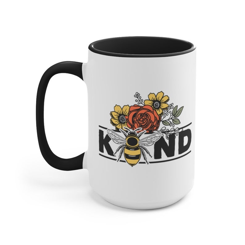 Op de afbeelding: Een witte keramische mok met een zwarte rand en handvat. De mok heeft een bij met gele en zwarte strepen, omringd door bloemen in tinten rood, geel en oranje. De tekst "KIND" is in zwarte letters boven de bij gedrukt.