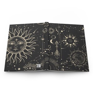 May include: An open book with a black cover and a white design of celestial bodies, including a sun, moon, stars, and constellations. The design is repeated on both pages of the book.