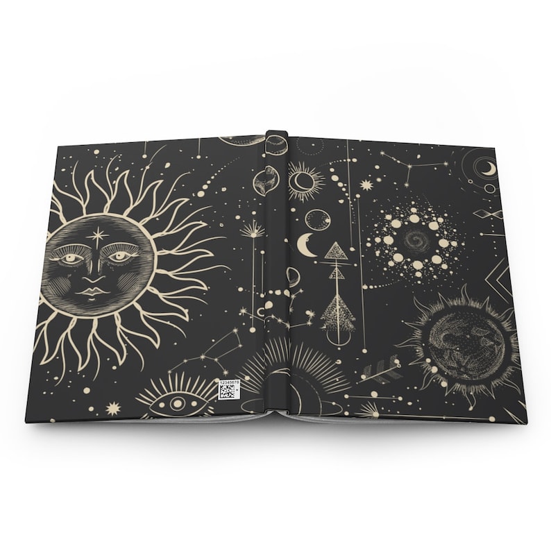 May include: An open book with a black cover and a white design of celestial bodies, including a sun, moon, stars, and constellations. The design is repeated on both pages of the book.