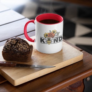 Op de afbeelding: Witte keramische mok met een rode rand en handvat. De mok heeft een zwarte en gele bij met bloemen en het woord "KIND" erop gedrukt. De mok is gevuld met koffie en er stijgt stoom op uit de bovenkant. Een bruine muffin ligt op een houten snijplank naast de mok.