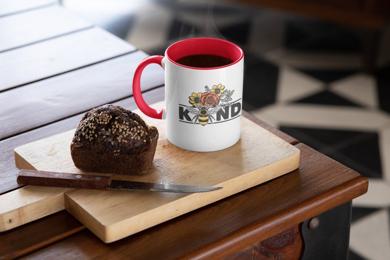 Op de afbeelding: Witte keramische mok met een rode rand en handvat. De mok heeft een zwarte en gele bij met bloemen en het woord "KIND" erop gedrukt. De mok is gevuld met koffie en er stijgt stoom op uit de bovenkant. Een bruine muffin ligt op een houten snijplank naast de mok.