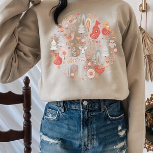 May include: A beige sweatshirt with a circular design featuring a deer, foxes, birds, and trees in a Scandinavian folk art style.