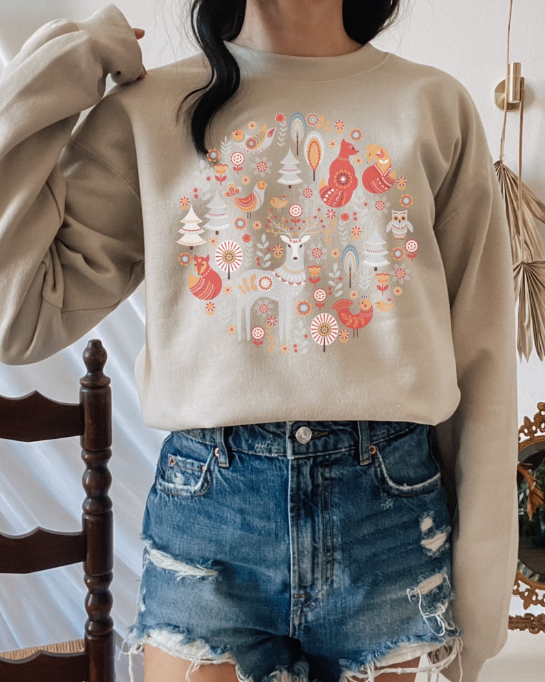 May include: A beige sweatshirt with a circular design featuring a deer, foxes, birds, and trees in a Scandinavian folk art style.
