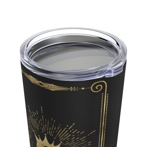 May include: A black stainless steel tumbler with a gold crown and sunburst design. The tumbler has a clear lid.