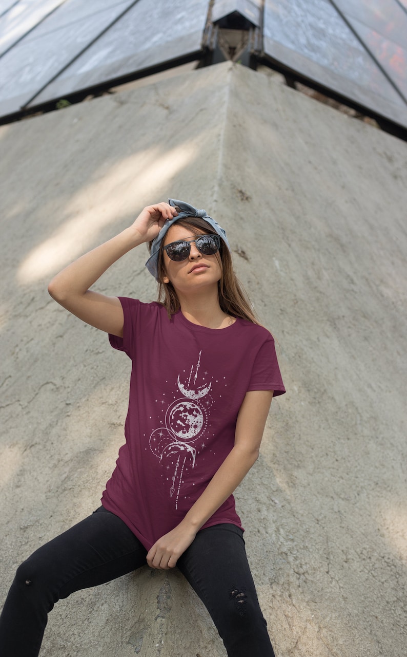 May include: A maroon t-shirt with a white graphic of three moons and stars. The graphic is centered on the shirt and has a minimalist design.