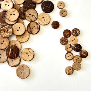 May include: A collection of brown and natural coconut shell buttons of various sizes. The buttons have two holes and are scattered on a white surface.