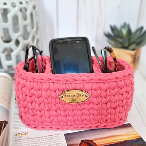 Puede incluir: Un organizador de crochet rosa con dos compartimentos para guardar gafas y un tel&eacute;fono. El organizador tiene una etiqueta de madera que dice "Farmhouse Stitches ByK".
