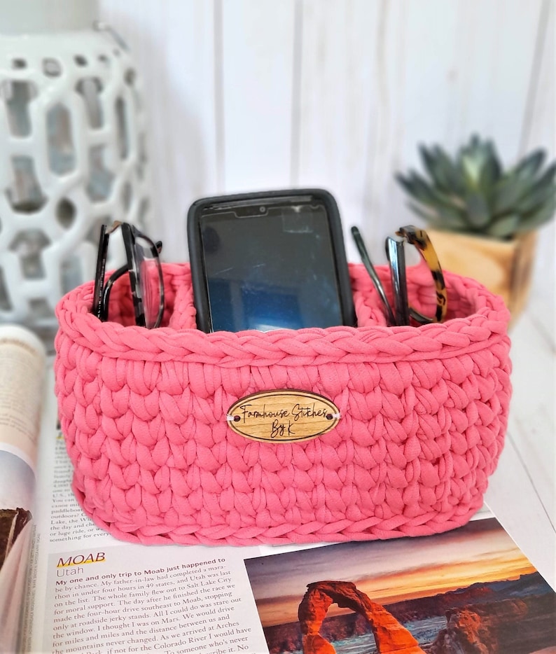 Puede incluir: Un organizador de crochet rosa con dos compartimentos para guardar gafas y un tel&eacute;fono. El organizador tiene una etiqueta de madera que dice "Farmhouse Stitches ByK".