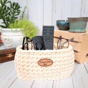 Puede incluir: Una cesta de crochet de color crema con una etiqueta de madera que dice "Farmhouse Stitchers BK" contiene dos mandos a distancia negros y un par de gafas.