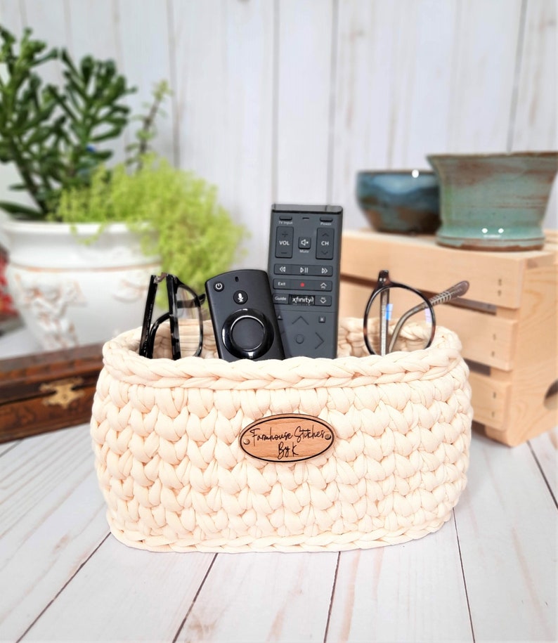 Puede incluir: Una cesta de crochet de color crema con una etiqueta de madera que dice "Farmhouse Stitchers BK" contiene dos mandos a distancia negros y un par de gafas.