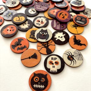 May include: A collection of 25 round wooden buttons with various Halloween-themed designs. The buttons feature images of pumpkins, ghosts, bats, owls, spiders, skulls, and other spooky elements. The buttons are painted in orange, black, white, and purple.