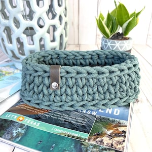 May include: A rectangular, teal-colored crochet basket with a brown leather strap and silver button closure. The basket is sitting on a magazine with the text "LET'S TEXAS" and "TRAVELTEXAS.COM".