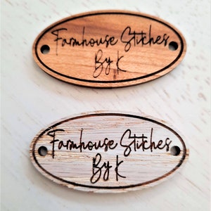 Puede incluir: Dos etiquetas ovaladas de madera con agujeros para una cuerda. La etiqueta superior es de madera marr&oacute;n claro con el texto "Farmhouse Stitches By K" quemado en la madera. La etiqueta inferior es de madera blanqueada con el mismo texto quemado en la madera.