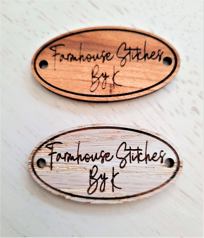 Puede incluir: Dos etiquetas ovaladas de madera con agujeros para una cuerda. La etiqueta superior es de madera marr&oacute;n claro con el texto "Farmhouse Stitches By K" quemado en la madera. La etiqueta inferior es de madera blanqueada con el mismo texto quemado en la madera.