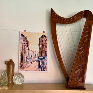 May include: A watercolor painting of a city street with a tall building in the background. The painting is framed and hanging on a wall next to a wooden harp.