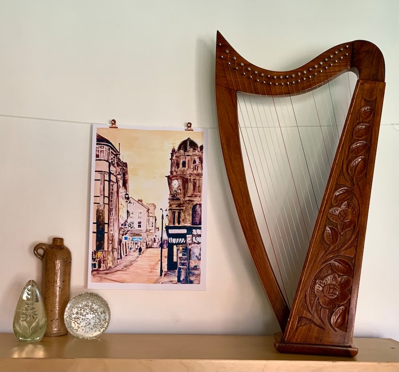 May include: A watercolor painting of a city street with a tall building in the background. The painting is framed and hanging on a wall next to a wooden harp.