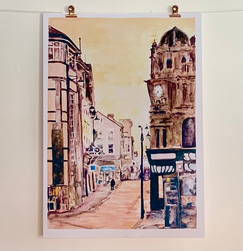 May include: A watercolor painting of a street scene with a clock tower in the background. The buildings are painted in shades of brown and yellow, and the street is empty except for a single figure walking in the distance.