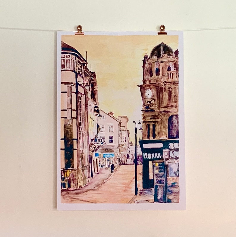 May include: A watercolor painting of a city street scene with a clock tower in the background. The buildings are painted in shades of brown and yellow, and the street is empty except for a single person walking in the distance.