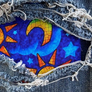 May include: Close-up of a denim fabric with a patch featuring a celestial design. The patch showcases a blue background with stars, a crescent moon, and sun-like shapes in yellow and orange. The denim has frayed edges.