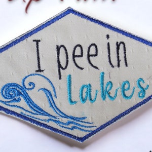 Puede incluir: Un parche hexagonal blanco con costuras azules alrededor de los bordes. El parche tiene bordado de olas azules y turquesa y el texto "I pee in lakes".