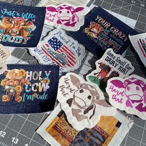 May include: A collection of embroidered patches with cow-themed designs. The patches feature phrases like "Holy Cow I'm cute" and "Just a little moody" with cow illustrations and floral accents. Some patches have an American flag design.