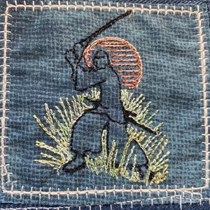 May include: Embroidered patch on denim fabric depicting a figure in a field of grass with a sword raised, a red sun behind them.