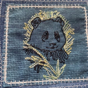 May include: A black and white embroidered panda bear with green and yellow flowers on a blue denim background.