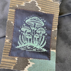 May include: A fabric patch featuring a cluster of mushrooms in a light green color, stitched onto a navy blue square. The square is sewn onto a patterned fabric with brown, tan, and teal colors. The patch is likely for clothing or accessories.
