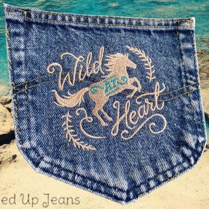 May include: A denim jean pocket with embroidered text that reads "Wild at Heart" with a stylized horse design in the center. The horse is a light brown color and the text is a light pink color.