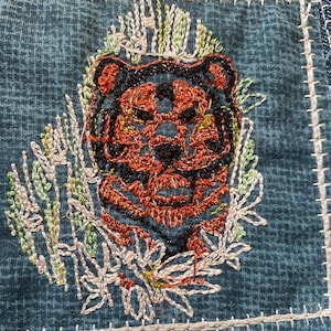 May include: Embroidered patch featuring a tiger's face in orange and black with a green and white floral design around it. The patch is sewn onto a blue denim fabric.