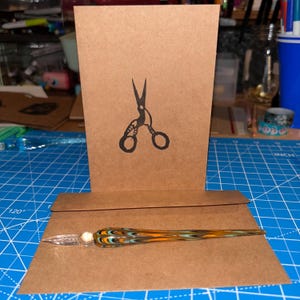 May include: A brown card with a black silhouette of a pair of scissors. The card is sitting on top of a brown envelope with a colorful glass dip pen on top of the envelope.
