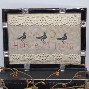 May include: A rectangular cross-stitch sampler featuring three black birds, a yellow crescent moon, and three small stars on beige fabric. The text "Harvest Moon" is stitched in pink. The piece is framed in a black border with mirrored accents.