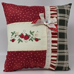 May include: Square cushion with a patchwork design. The cushion features red polka dot fabric, a cross-stitched strawberry design, and a green plaid pattern. It is embellished with lace, ribbons, and a button.
