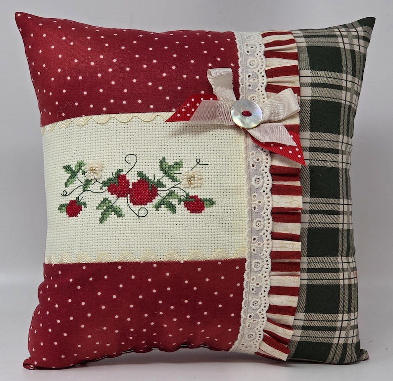 May include: Square cushion with a patchwork design. The cushion features red polka dot fabric, a cross-stitched strawberry design, and a green plaid pattern. It is embellished with lace, ribbons, and a button.