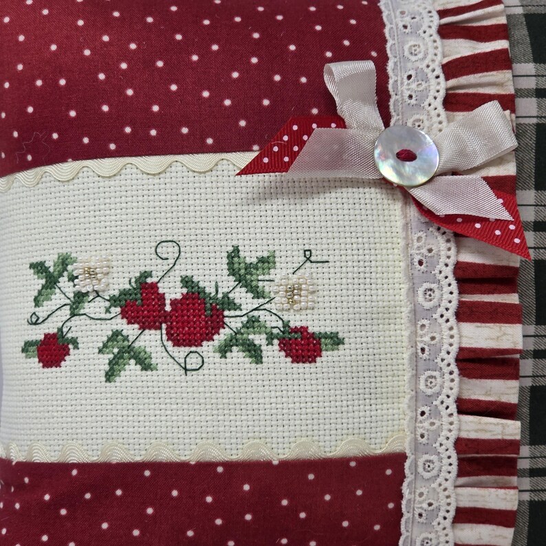 May include: A cushion with a red and white polka dot pattern and a cross-stitched strawberry design. It has white lace trim, a ribbon bow, and a button closure. The color scheme is red, white, and green. The cushion is square-shaped.