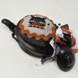 May include: A black ceramic teapot pincushion featuring a cross-stitched cat design on a light gray fabric insert, bordered by orange rickrack trim. The teapot is adorned with a white button and layered ribbons in gray, white, and orange.
