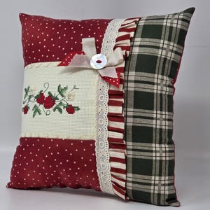 May include: Square decorative cushion with a patchwork design. The cushion features sections of red polka dots, cross-stitched strawberries, and green and black plaid. A white lace trim and a bow with a button accent the design.