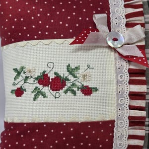May include: A cushion with a red and white polka dot pattern and a cross-stitched strawberry design. The cushion features a white lace trim, a ribbon, and a button closure. The colour scheme is red, white, and green.